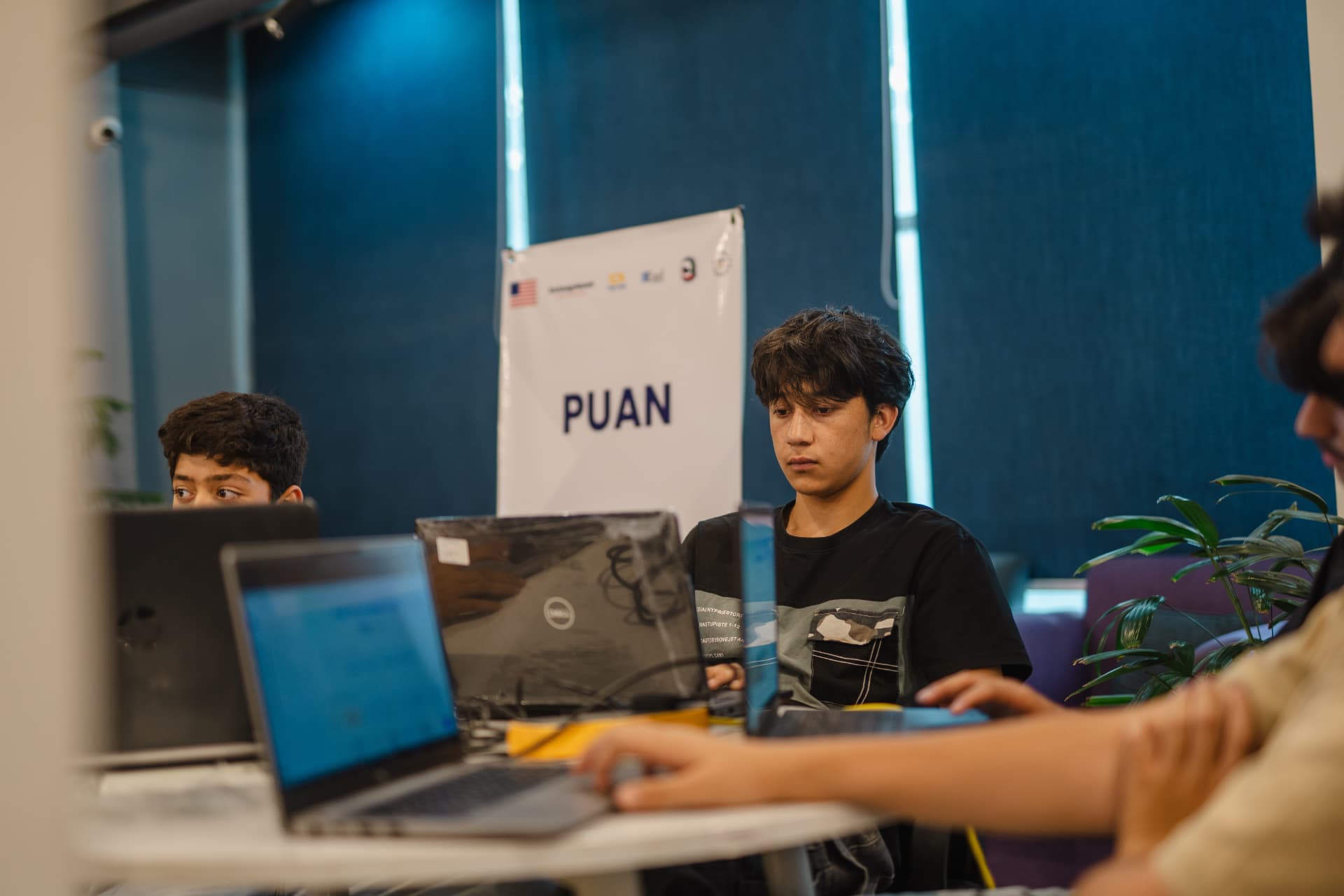 Two young participants building projects on laptops beside the PUAN banner